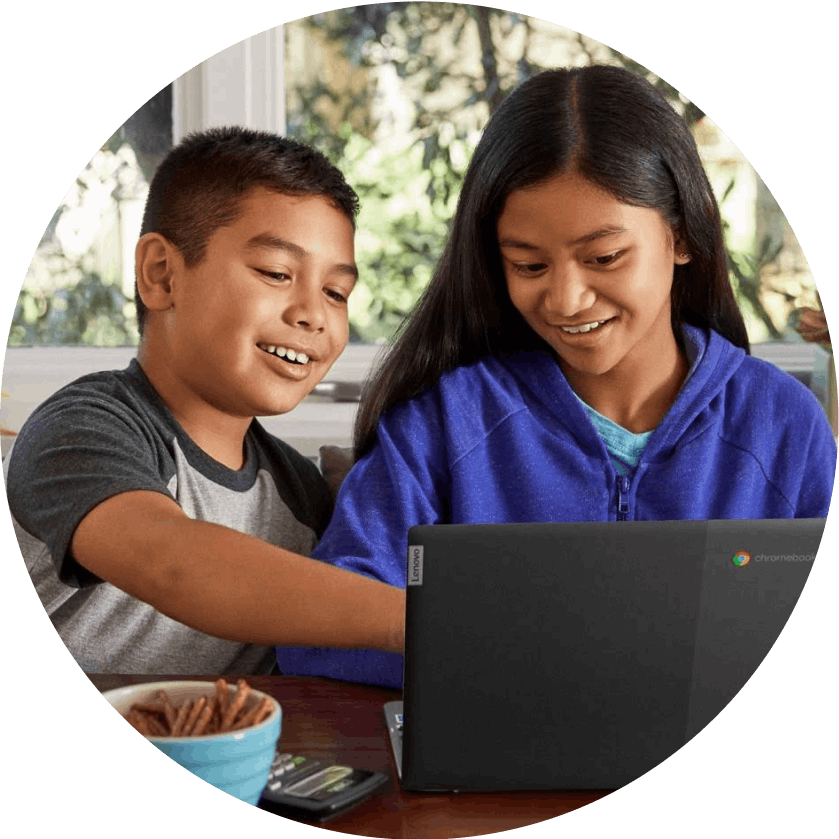 Two smiling children sitting in front of a Chromebook, with one pointing at the screen